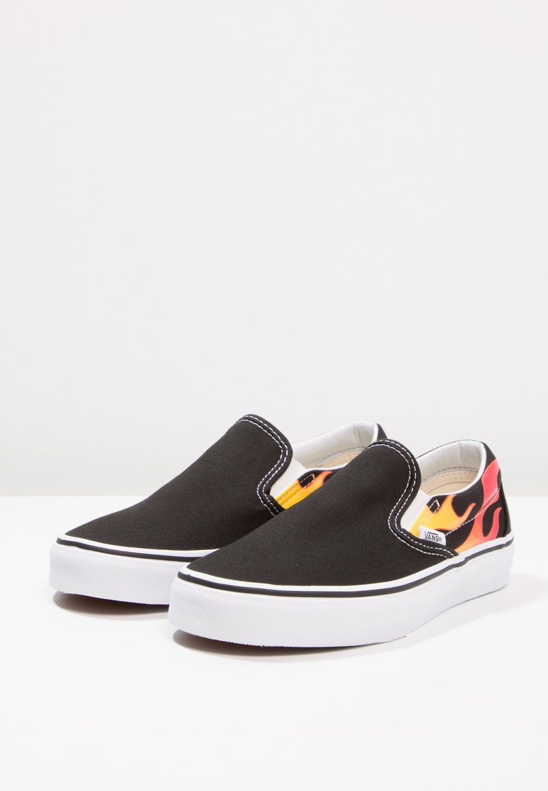 Zapatos slip-on de lienzo negro con suela blanca, que presentan un diseño de llamas en naranja y rojo en el talón, junto con costuras blancas contrastantes.