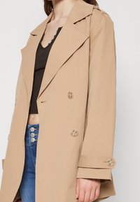Beige dubbelzijdige trenchcoat van zachte stof, met grote knopen, brede revers en een losse pasvorm, gedragen over een zwart top.