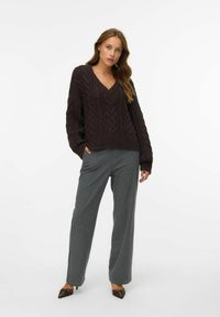 Pull en tricot brun foncé avec un col en V et un motif en torsades, associé à un pantalon à rayures gris clair et des chaussures à bout pointu marron.