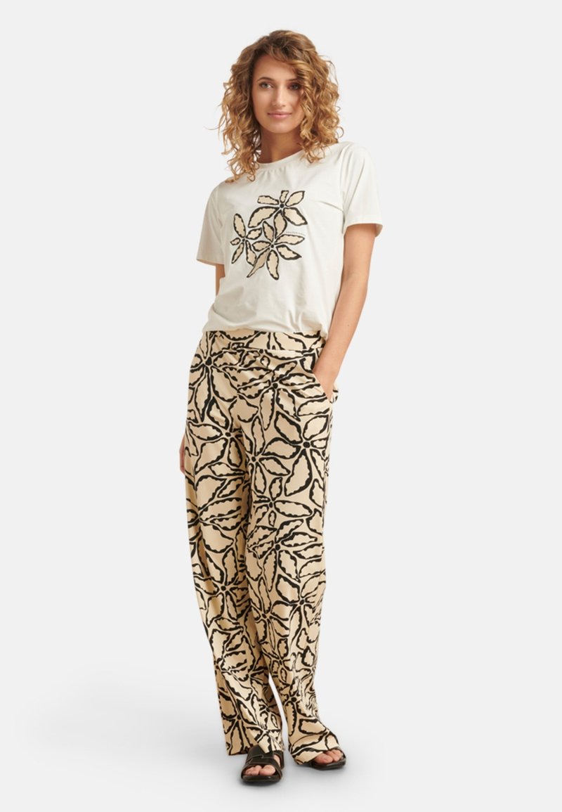 Crème t-shirt met zwart bloemenpatroon; gecombineerd met beige broek met zwart bladerenpatroon; model draagt zwarte sandalen.