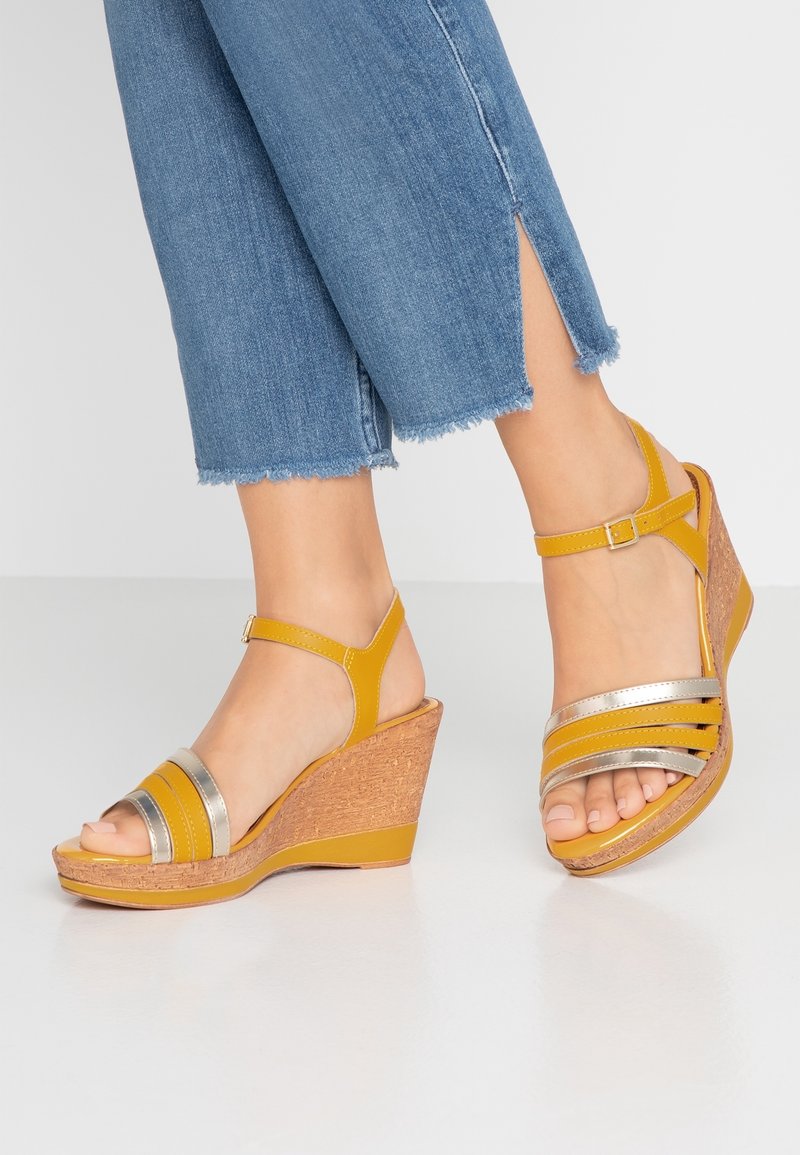 Sandales compensées jaunes avec accents argentés rayés, texture semblable à du liège sur la semelle compensée, bride à la cheville avec boucle, et design à bout ouvert.