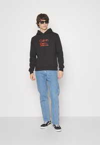 Black hooded sweatshirt with red "Calvin Klein" logo, paired with light blue jeans and black sneakers, standing against a white background.