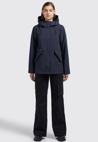Giacca impermeabile blu navy con cappuccio, zip frontale e due ampie tasche laterali; abbinata a pantaloni cargo neri e stivali neri.