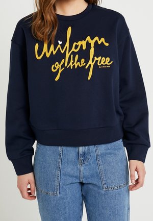 Femme portant un sweat-shirt bleu marine avec un texte jaune "Uniform of the free" et un jean taille haute bleu clair avec de grandes poches avant.