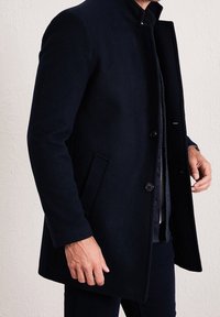 Tmavomodrý vlněný kabát s vysokým límcem, zapínáním na přední knoflíky, bočními kapsami a střihaným střihem, který se vyznačuje hladkou texturou a moderním designem.