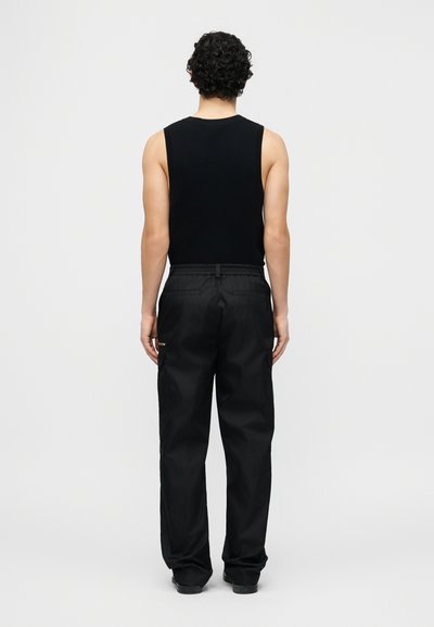 Άνδρας με σγουρά μαλλιά, φορώντας μαύρο αμάνικο τοπ και φαρδιά μαύρη παντελόνια, στέκεται αντικρίζοντας ένα απλό ανοιχτό γκρι φόντο.