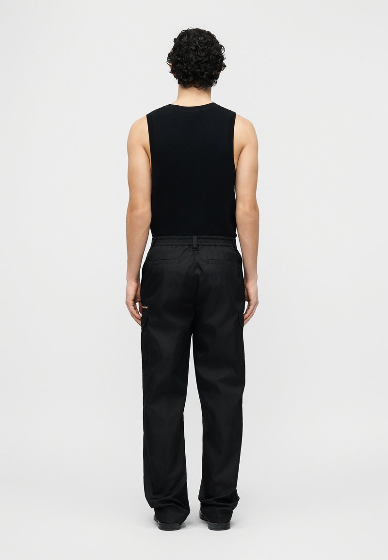 Homme aux cheveux bouclés portant un haut noir sans manches et un pantalon noir ample, debout face à un fond gris clair uni.