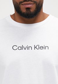 Fehér pamut póló, kerek nyakkal, a mellkason vastag, sötét betűkkel nyomtatva a "Calvin Klein" felirat. Látható a puha textúra.