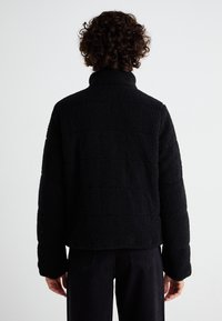 Jachetă din polar neagră, cu guler înalt și țesătură texturată. Are un design scurt și este purtată cu pantaloni negri, evidențiind contururi netede.