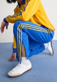 Chándal que incluye una parte superior amarilla con rayas azules y pantalones azules a juego. Zapatillas blancas con acentos grises. Textura de tela suave.