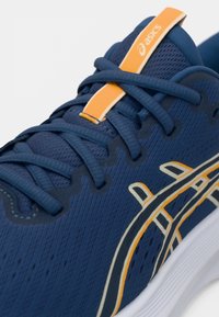 Μπλε αθλητικό παπούτσι για τρέξιμο με πορτοκαλί λεπτομέρειες, υφασμάτινο επάνω μέρος με υφή, στρογγυλή μύτη, επίπεδα κορδόνια και ορατό λογότυπο με καμπυλωτή σχεδίαση.