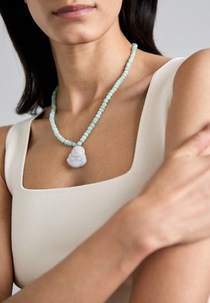 Una collana di perline con pietre di colore blu-verde chiaro e un pendente bianco intagliato a forma di figura seduta, indossata con una maglietta crema.