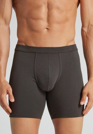 Mandligt overkrop iført tætsiddende mørkegrå boxershorts med synlige sømdetaljer mod en neutral baggrund.
