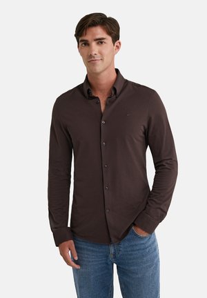 Hombre con camisa de botones marrón y jeans azules, de pie con una mano en el bolsillo contra un fondo claro y liso.