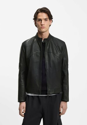 Joven de pie, vistiendo una chaqueta de cuero negra con cremallera sobre pantalones oscuros y una camiseta negra, frente a un fondo blanco liso.