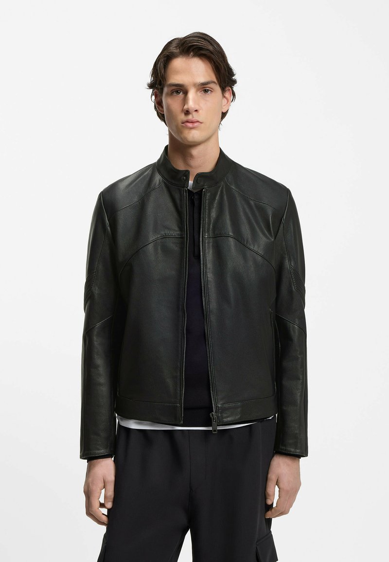 Junger Mann steht da, trägt eine schwarze Lederjacke mit Reißverschluss über einer dunklen Hose und einem schwarzen Hemd vor einem schlichten weißen Hintergrund.