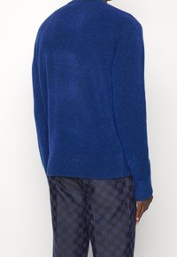 Blå stickad tröja med långa ärmar, rund hals och en strukturerad yta. Matchad med mönstrade mörka byxor med en repetitiv logotypdesign.