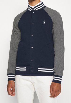 Person trägt eine marineblaue und graue Collegejacke mit weiß gestreiftem Kragen, Bündchen und Bund, kombiniert mit weißen Hosen.