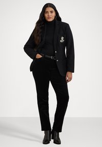 Črni blazer z odrezanim ovratnikom, ki ima srebrne gumbe in vezeno emblemo na žepu. Obut je nad črnim polo in hlačami.