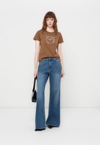Brown cotton t-shirt with a circular graphic design, paired with wide-leg blue denim jeans. Model holding a black handbag.