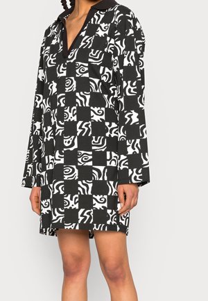 Robe à manches longues en noir et blanc avec un motif damier de tourbillons abstraits, portée par une personne aux jambes et mains visibles, sur un fond uni.