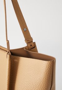 bruine leren totebag met een gestructureerde afwerking, voorzien van een strakke vorm en een platte schouderband die is bevestigd met een metalen accent.