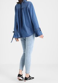 Tummansinisestä farkkupaidasta, jossa on kellotetut hihat ja solmukuvio. Yhdistetty vaaleansinihin slim-fit-farkkuihin ja mustiin lenkkareihin. Takakuvassa, yksinkertainen tausta.