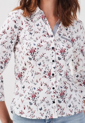 Femme portant une chemise blanche boutonnée avec un imprimé floral rouge et des papillons gris, manches longues, poche poitrine bordée de gris et jean bleu.