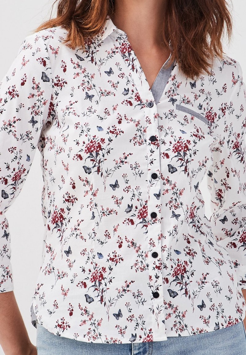 Femme portant une chemise blanche boutonnée avec un imprimé floral rouge et des papillons gris, manches longues, poche poitrine bordée de gris et jean bleu.