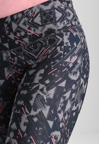 Legginsy w czarno-szarym wzorze mają geometryczne desenie z różowymi akcentami. Wykonane z elastycznego materiału, mają dopasowany, wysokozasłonięty krój.