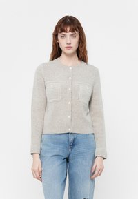 Lys grå strikket cardigan med rund halsutringning, knappelukking og to brystlommer. Har ribbestrikkede mansjetter og en myk tekstur.