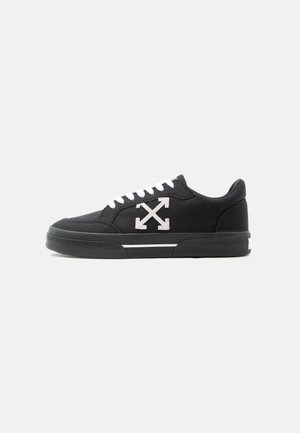 Baskets basses noires avec lacets blancs, semelle blanche rayée et logo flèche croisée blanche sur le côté.