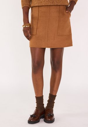 Bruine wollen mini-jurk met twee voorzakken, gecombineerd met bruine loafers en geribde bruine sokken. De textuur lijkt zacht en gezellig.