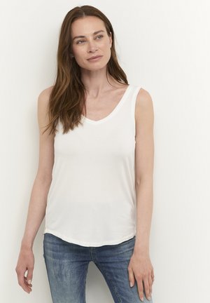 Femme aux longs cheveux bruns portant un haut blanc sans manches et un jean bleu, debout devant un fond blanc uni.