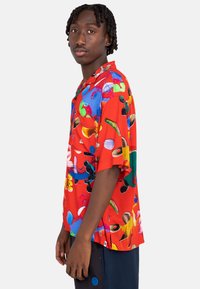Short-sleeve red shirt featuring a vibrant, abstract pattern with blue, green, and yellow shapes. Casual fit with a camp collar design.