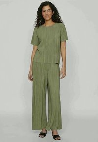 Conjunto de duas peças em verde-oliva, apresentando uma blusa de manga curta com um corte solto e calças de perna larga. O tecido possui pregas verticais e uma textura suave.