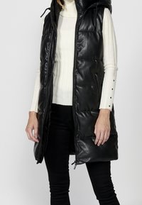 Femme portant une veste en cuir noire matelassée sans manches sur un pull à col roulé blanc côtelé et un pantalon noir.