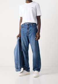 Witte katoenen T-shirt met "HUGO" logo, gecombineerd met losvallende blauwe spijkerbroeken. De persoon houdt een spijkerjack vast en draagt witte sneakers.