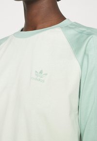 Světle zelená a krémová košile s dlouhým rukávem, raglánovými rukávy a malým vyšitým logem Adidas na hrudi. Materiál z bavlněné směsi.