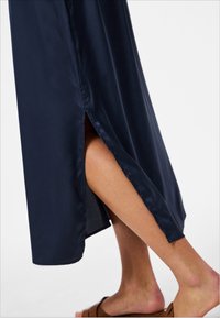 Jambe en sandale mule marron en train de marcher, portant une jupe bleu marine fendue sur le côté laissant apparaître le mollet inférieur et la cheville.