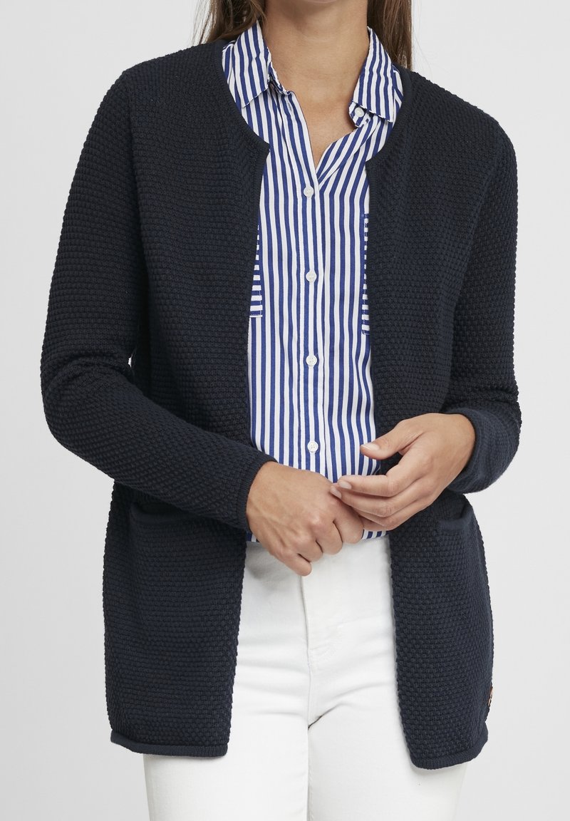 Marineblau gestrickte Strickjacke mit offenem Vorderteil, gerippten Bündchen und Taschen, getragen über einem blau-weiß gestreiften Hemd mit Knopfleiste.