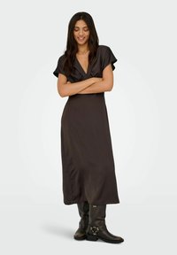 Ruda satino ilga suknelė su gilia V formos iškirpte ir trumpomis rankovėmis, pasižyminti sklandžia tekstūra ir plačia siluetu, derinama su iki kelių siekiančiais juodais batais.