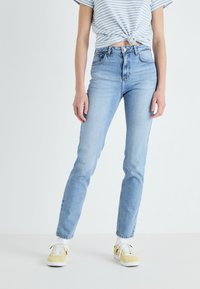 Jeans de cintura alta azul clara em ajuste slim, com design de bolso frontal e acabamento desbotado. Combinados com calçado amarelo e uma blusa às riscas.