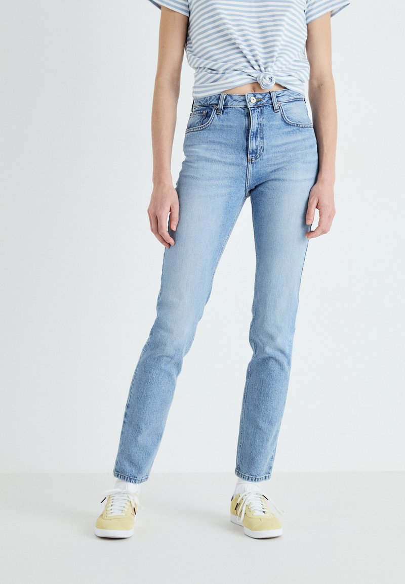 Jeans de cintura alta azul clara em ajuste slim, com design de bolso frontal e acabamento desbotado. Combinados com calçado amarelo e uma blusa às riscas.