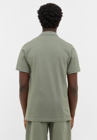 Khaki krótki rękaw polo z wyraźnym kołnierzykiem, gładkim bawełnianym materiałem i podwiniętymi mankietami. Widok z tyłu pokazuje luźny krój i minimalne detale.