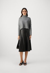 Grijze cropped trui met een geribbelde structuur, gecombineerd met een zwarte leren midi-rok. Model houdt een zwarte handtas vast en draagt zwarte open teen schoenen.