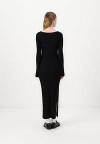 Abito a costine nero con maniche lunghe, spacco laterale e scollatura ampia. Il modello indossa scarpe nere e calzini bianchi. Sfondo bianco semplice.