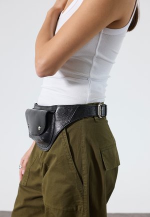 Donna che indossa pantaloni cargo verde oliva e canotta bianca con marsupio in pelle nera intorno alla vita su sfondo neutro.