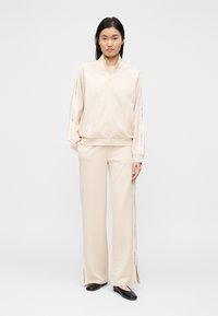 Bež trenirka z zadrgo na ovratniku, bočnimi razporkami na hlačah in potiskanimi poudarki po rokavih. Gladek tekstura tkanine. Črne ravne čevlje.