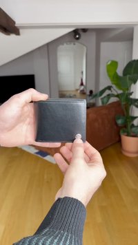 Černá kožená peněženka držená v ruce, s hladkou texturou a zaobleným tlačítkem. V pozadí je obývací prostor s rostlinami.